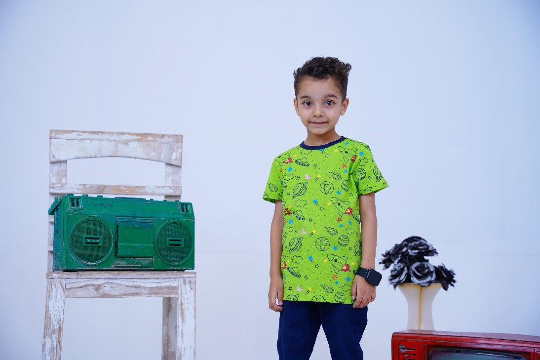 A child stands in front of a white chair with a green boombox, wearing a bright green T-shirt with a space-themed pattern and blue pants. A black and white floral arrangement and a vintage red TV are in the background.