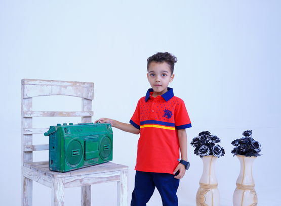A young boy in a red polo shirt and blue pants stands next to a green boombox on a wooden chair, with two vases of black flowers in the background