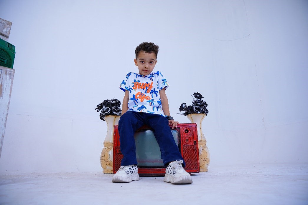 A young child sitting on a red vintage TV, wearing a colorful "Surf Life" shirt, blue pants, and white sneakers, with black floral arrangements and a green speaker in the background