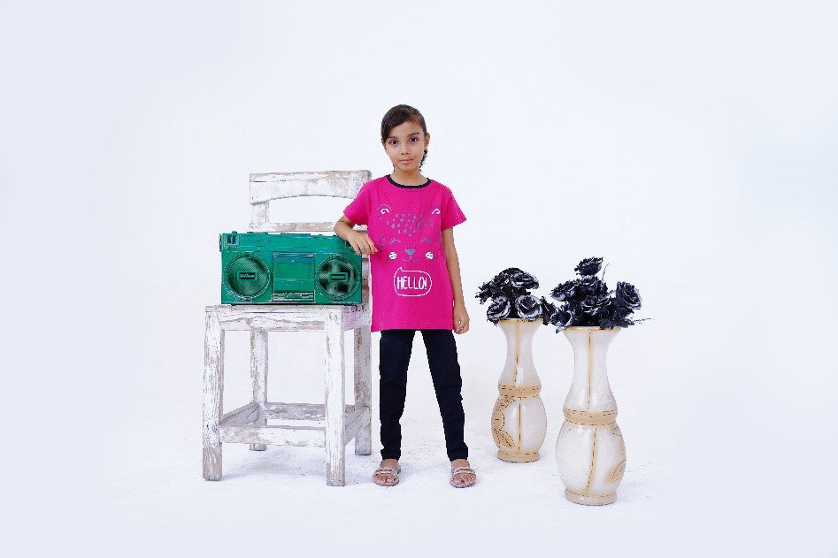 A young girl in a pink shirt and black pants stands next to a wooden chair with a green boombox, with two vases of black flowers beside he